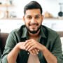 Portrait of confident attractive indian or arabian man, in casual stylish clothes, sitting at home in cozy living room on the background of the kitchen, looks at camera, smiling friendly