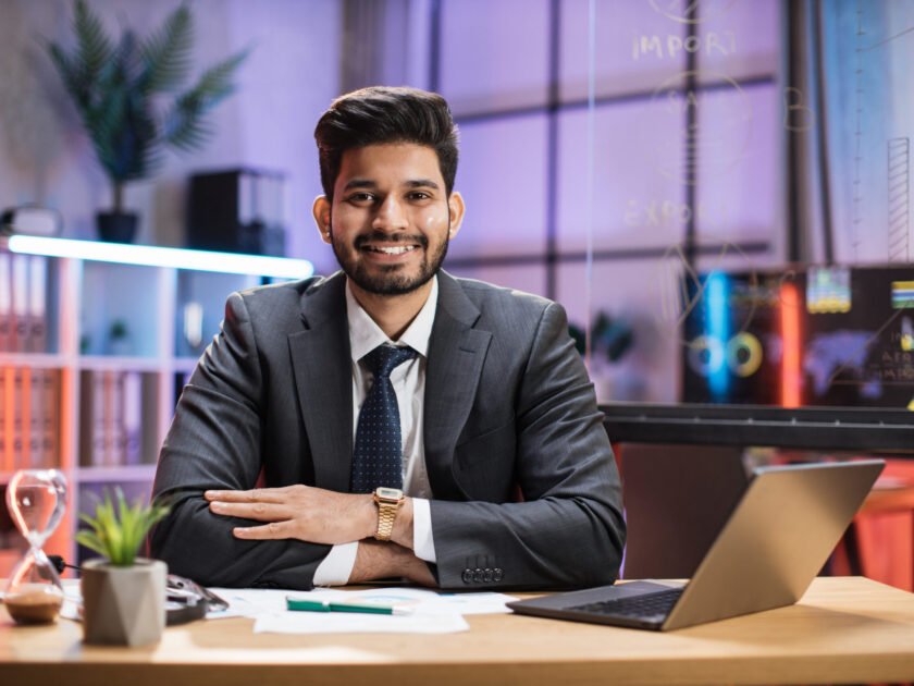 Close up portrait of smiling confident indian financial expert office worker sitting at table in formal suit working with laptop in evening modern office.