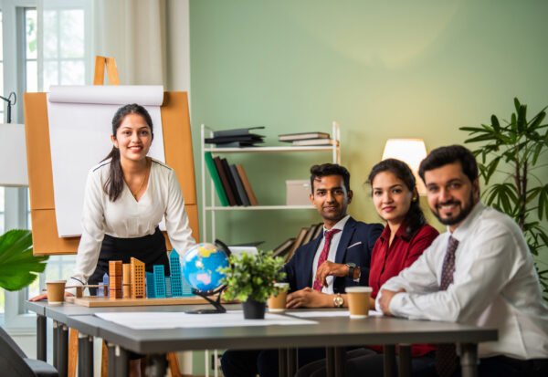 Asian Indian leader in 30s coaching interested business people, giving educational workshop presentation in office. Concentrated employees listening to professional coach at workshop.