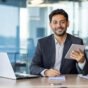 Portrait of young businessman with tablet computer inside office, hispanic man smiling and looking at camera, working with laptop, using app on tablet.