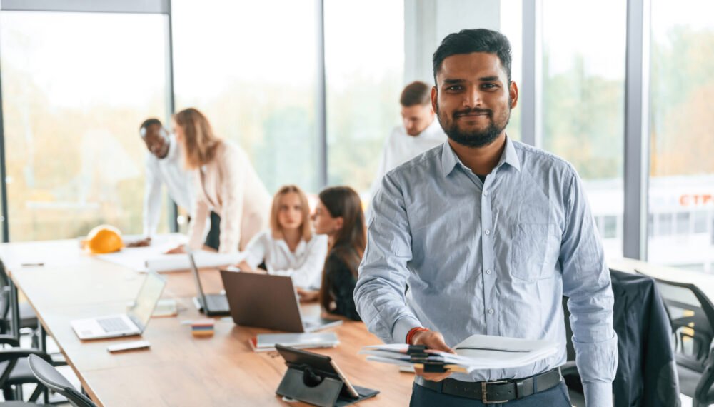 Indian man is standing in front of colleagues that are using laptop. Group of office workers are together indoors.