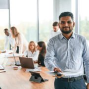 Indian man is standing in front of colleagues that are using laptop. Group of office workers are together indoors.